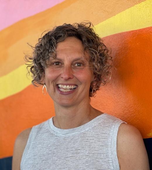 Person with short curls and wearing a white top smiling again a wall painted with pink, orange, and yellow hues.