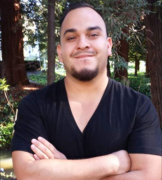 Photo of Jose Manuel smiling at the camera with his arms crossed in front of his chest; they are wearing a black shirt and standing in front of some trees