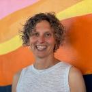 Person with short curls and wearing a white top smiling again a wall painted with pink, orange, and yellow hues.